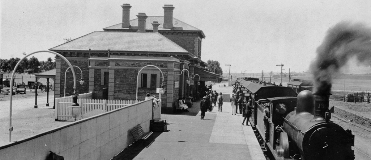 Image: A railway station. A train is on the platform to the right, with smoke coming out of the front. People are boarding the train. To the left is the station building, made of brick