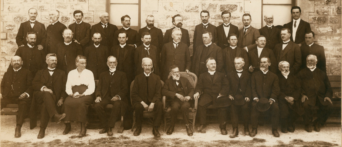 Image: Black and white group portrait. Three rows of men in suits pose against a stone building. There is one woman in the photograph. She is seated in the front row, third from left.