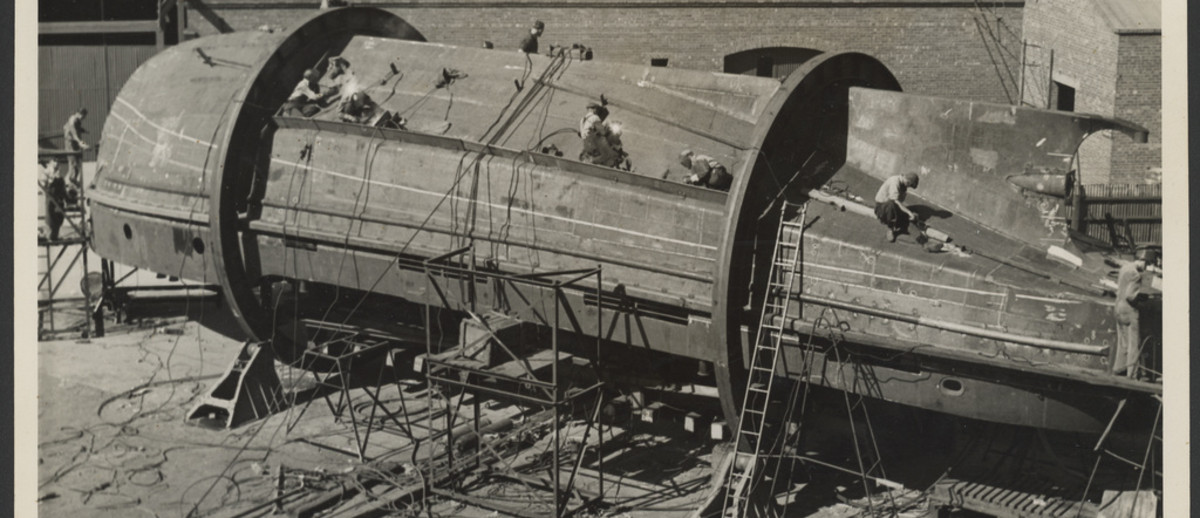 Image: The body of a tug boat upside down in the process of being constructed, numerous people, ropes and ladders surround the boat