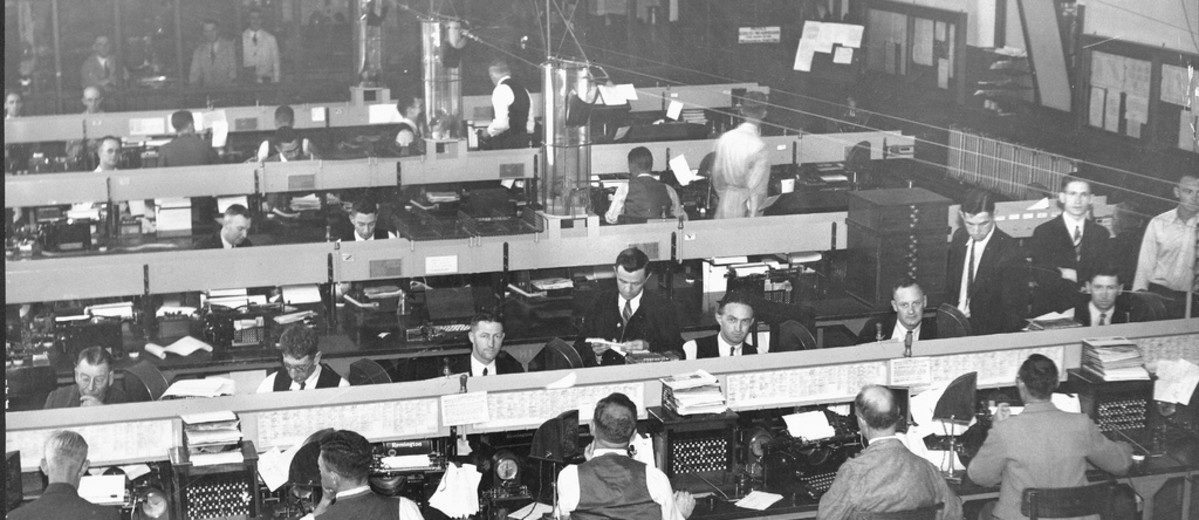 Image: The inside of a telegraph office. Men sit at rows of desks with telegraph machines