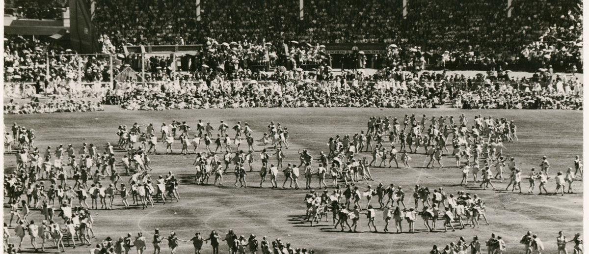 Image: An oval and grandstands filled with people, children dance on the oval in circular formations