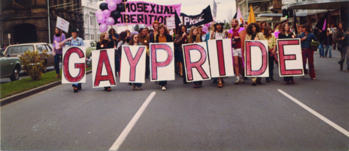 Image: men and women in 1970s era clothes march down a city street with pink and black balloons and signs reading "gay pride" and "homosexual liberation"