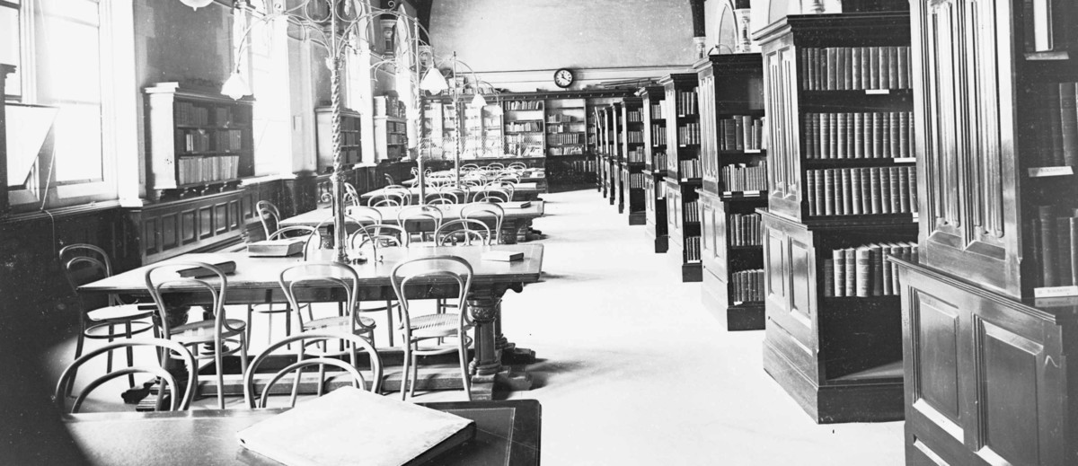 Image: A long, well-lit room containing several shelves of books on one side and six reading tables on the other