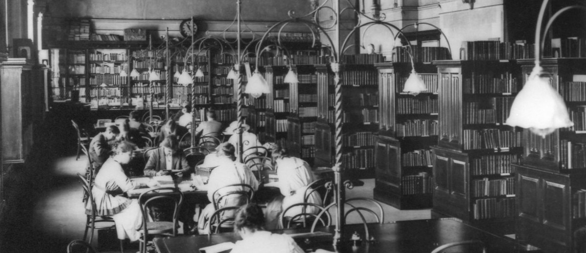 Image: male and female students sit at tables in a library with a vaulted ceiling 