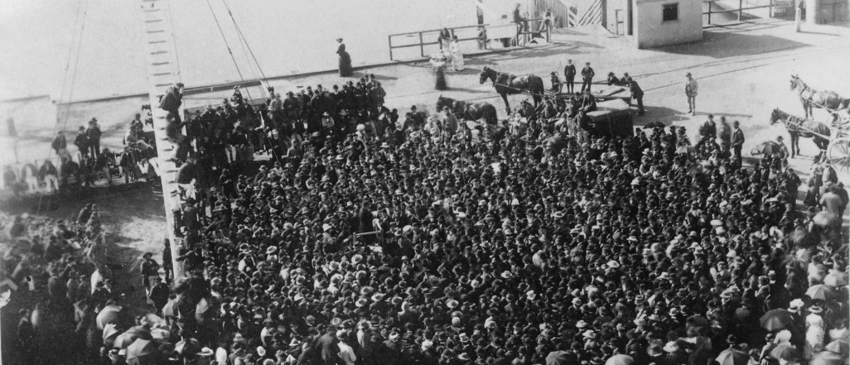 Image: a large crowd of workers gather for a strike at a wharf, the ocean can be seen in the background