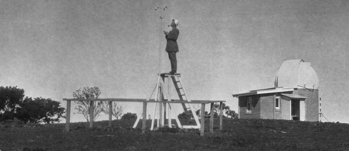 Image: A man, smoking a pipe, stands on a ladder collecting data at an outside, rural weather observatory