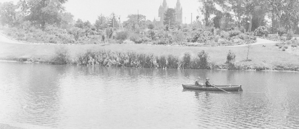 Image: Two men sitting in a row boat on a River, with a park and Cathedral visible in the background 