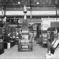 Image: a number of displays in glass cabinets fill a large hall. In the centre of the photograph a display of varnishes is topped by a pyramid. To the left a man stands in a uniform of a dark jacket and white pants and hat.