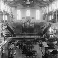 Image: a pipe organ takes pride of place at the far end of a large hall with a hammer beam roof. In the foreground men and women in black sit on rows of chairs surrounded by glass display cabinets. 