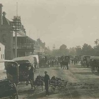 Image: A boy holding two hand-carts stands to the side of a busy street surrounded by horse-drawn carts. A tram line runs down the middle of the street, stone and brick buildings, one with a large balcony, line one side while the other fronts parklands.
