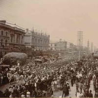 Image: a large crowd of people watch a contingent of soldiers in late 19th century military uniforms march down a wide city street led by four men on horseback