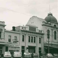 Image: a row of 1950s era cars are parked on a street outside of a row of two and three storey commercial buildings including a hotel with a verandah and balcony and a bank with a domed roof and flag pole.