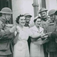 Image: Two women in white dresses hand cigarettes to a group of five men in military uniforms and helmets