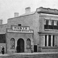 Image: a simple two storey building with an arched door to one side, a balconet and a small parapet stands next to a single storey building with a central arched door flanked by arched windows and a parapet sign reading Hunt's Labour Office