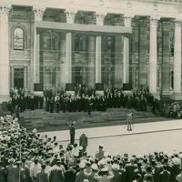Image: a man stands in front of a wide staircase leading up to a grand building with columns, photographing the men who are arranged upon it. A large crowd surrounds him.