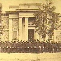 Image: a large group of men in 1860s police uniforms stand in front of a two storey stone building with columns flanking its entrance. 