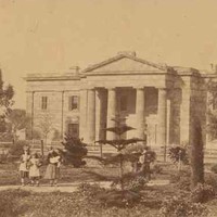 Image: three young girls walk through a garden in front of a two storey stone building with a portico featuring a triangular pediment supported by four columns