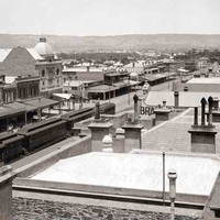 Image: a train is stopped in the centre of a city street which is lined with two and three storey buildings most of which have verandahs and one which features a large domed roof.