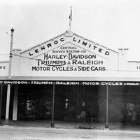 Image: a single storey shop with a verandah and triangular parapet with a painted sign reading "Lenroc Limited Central Service Station for Harley-Davidson, Triumph & Raleigh motor cycles & side cars". A flag flies from the peak of the parapet.