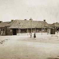 Image: Three girls in late 19th century dresses play in a wide dirt road while older women stand together outside stores with wooden shingled roofs and verandahs.  In the centre of the image a gas street light can be seen.