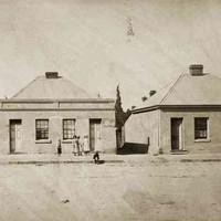 Image: a group of three pairs of semi-detached simple fronted cottages with a single door and window each and a central chimney rising from each roof. In front of the buildings stand women and children in late 19th century clothing. 