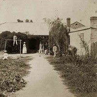 Image: A family (a man, woman, three boys and two girls) and their dog pose in an overgrown garden outside a single storey cottage with verandah. 