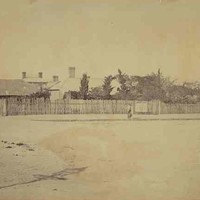 Image: fruit trees, tin roofs and brick chimneys rise above a paling fence bordering a dirt road. A number of people in late 19th century dress can be seen standing in front of the fence. 