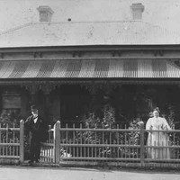 Image: a man and a woman stand behind a wooden fence in front of a bluestone cottage with tin roof and verandah. The man stands at the gate and wears a dark suit and bowler hat, the woman who is to the right of the photograph wears a white dress.