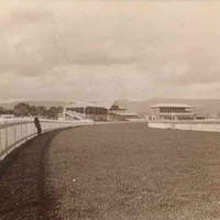 Image: A man leans against a high picket fence lining a curved racecourse. Two  grandstands can be seen in the distance.