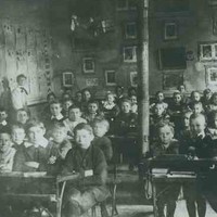 Image: a large group of boys and girls in early 20th century clothing pose sitting at their desks in a school room. In the background their teachers watch over them.