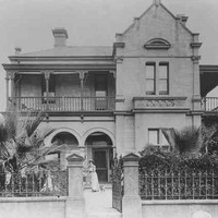 Image: A woman in a white dress and her small dog stand in front of a two storey house with a bay window, arched verandah and balconies. 