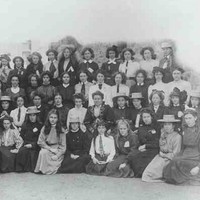 Image: a large group of girls in early 20th century dresses, some wearing straw hats, pose in four rows for this formal school photograph.