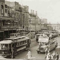 Image: a city street busy with pedestrians, electric trams and buses. In the left foreground is the Adelaide-Glenelg Railways Bus, a double decker with an open top, its passengers enjoying the sunshine. One of the buildings is in process of remodelling.