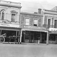 Image: a row of two and three storey commercial buildings with verandahs and/or balconies including the Crown and Sceptre Hotel, the Fish, Oyster and Grill Saloon, A.F. Shelton's Ham Shop and the King's Theatre. 