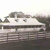 Image: Black and white photograph of homestead with outbuildings and a wooden fence in foreground