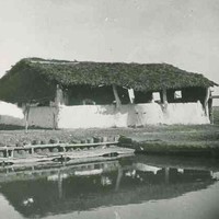 Image: A wooden framed building with a thatched roof and half height mud-brick walls stands behind a square edged pool in a barren field