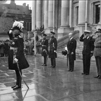 Image: a small group of men in military dress uniforms stand saluting on the pavement in front of a set of stone steps leading up to a building with columns. 