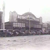 Image: a row of 1920s era cars are parked on a 45 degree angle facing a dirt road. Behind them is a single storey commercial building, part of a row of terraced shops, with a curved parapet and a sign which reads "Autocars Limited".