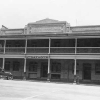 Image: A two storey hotel with balcony set within a terrace. 1920s era cars are parked on the street outside.