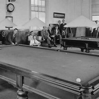 Image: a group of men sit or stand around two large billiard tables. 