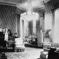 Image: late 19th century drawing room with high ceilings, a large chandeleir and heavily-patterned wallpaper and furniture