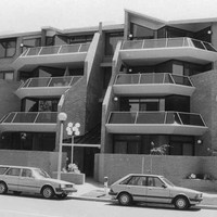Image: Three 1980s era cars are parked in front of four-storey 1980s "modern" brick townhouses with angular glass railed balconies.