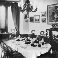 Image: The dining room of a large house in the 1890s. The room has a number of paintings on the wall, a bay window, and the table is set for eight. 
