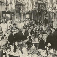 Image: a large group of men in dark suits are seated around long tables dressed with white tablecloths and large floral arrangements. Women in white dresses and hats stand in lines on either side of the room and streamers hang from the roof.