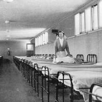 Image: a woman in a nurses uniform makes a bed halfway down a long row of beds. A second row lines the opposite wall.