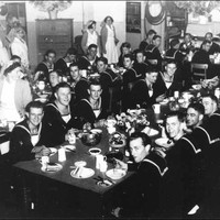 Image: A group of men in sailors uniforms sit around tables while women in white uniforms serve them food