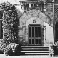 Image: Front entrance to large stone house. Stone steps lead up to the arched front door, flanked by two columns. To the left a window is completely surrounded by ivy and to the right is a verandah with decorative wooden fretwork.