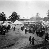 Image: a crowd of men and women in early 1870s clothing stand amongst tents and agricultural displays. In the background a large building can be seen.