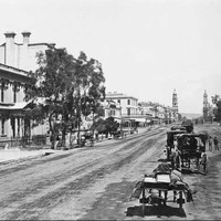 Image: Horses and buggies travel down a wide dirt road. In the distance two towers (the Town Hall and Post Office) can be seen on either side of the street. 
