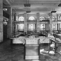 Image: the interior of a bank which is lined with marble and features a mezzanine balcony level, double height arched windows and square columns